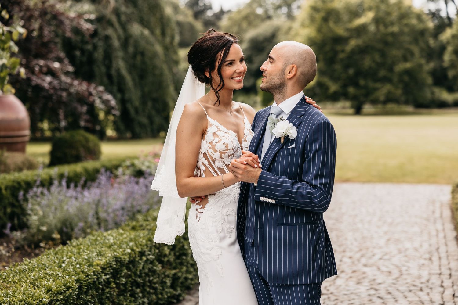 Smiling bride in lace gown and groom in navy pinstripe suit embrace in a lush garden.