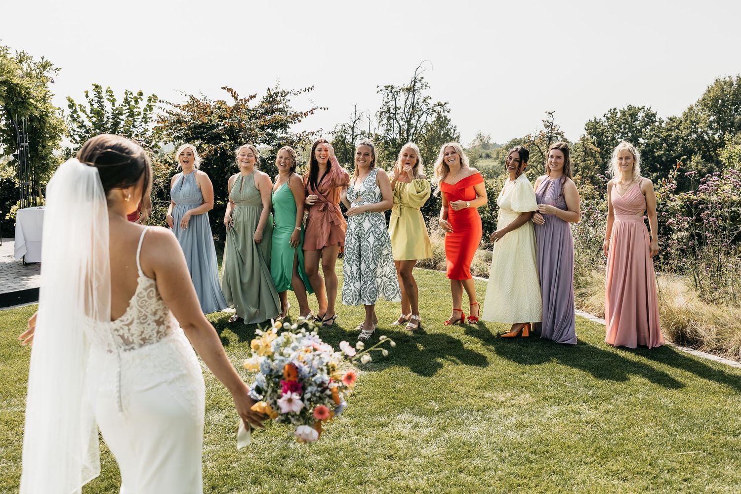 Bride holding colorful bouquet facing excited bridesmaids in rainbow of colorful dresses outdoors.