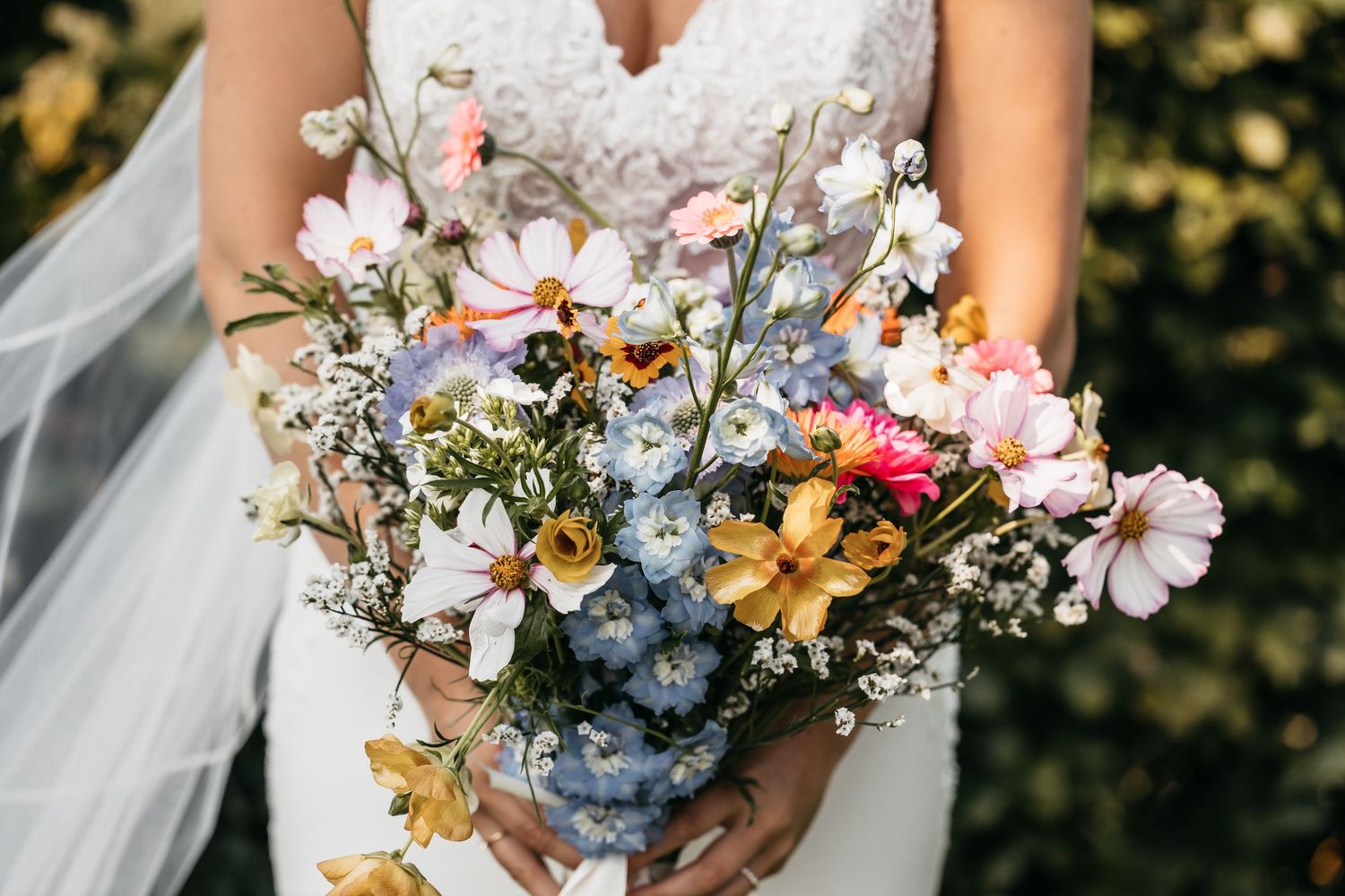 Bride holding lush colorful bouquet of cosmos, daisies, and wildflowers in white gown.