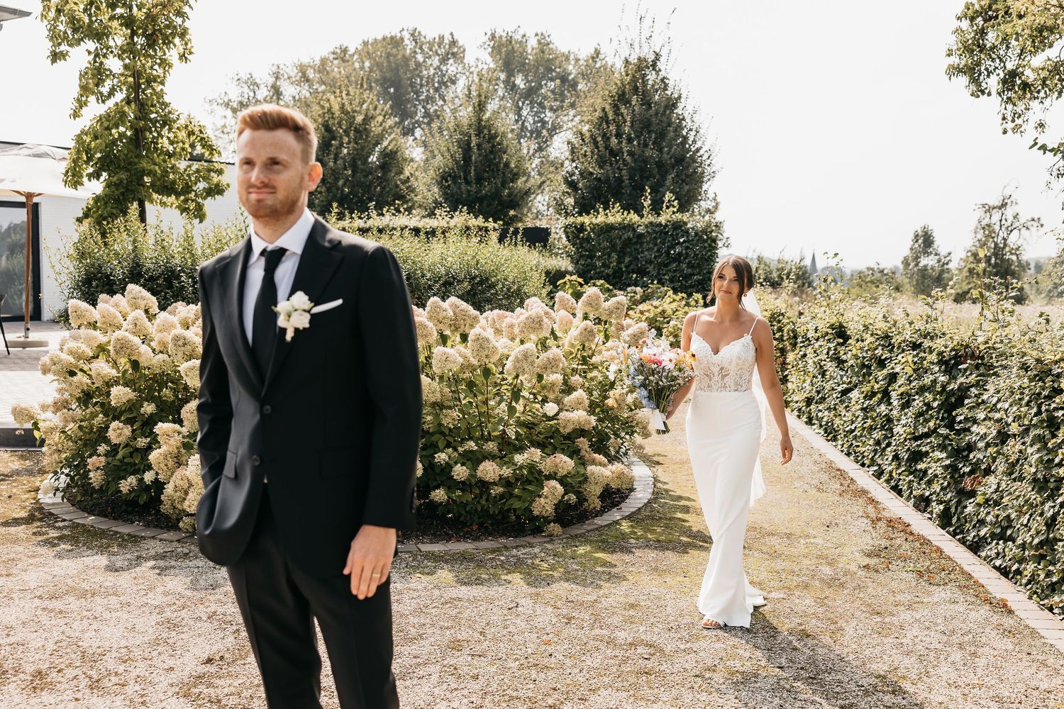 Groom in black suit stands as bride in white gown approaches on a garden path.