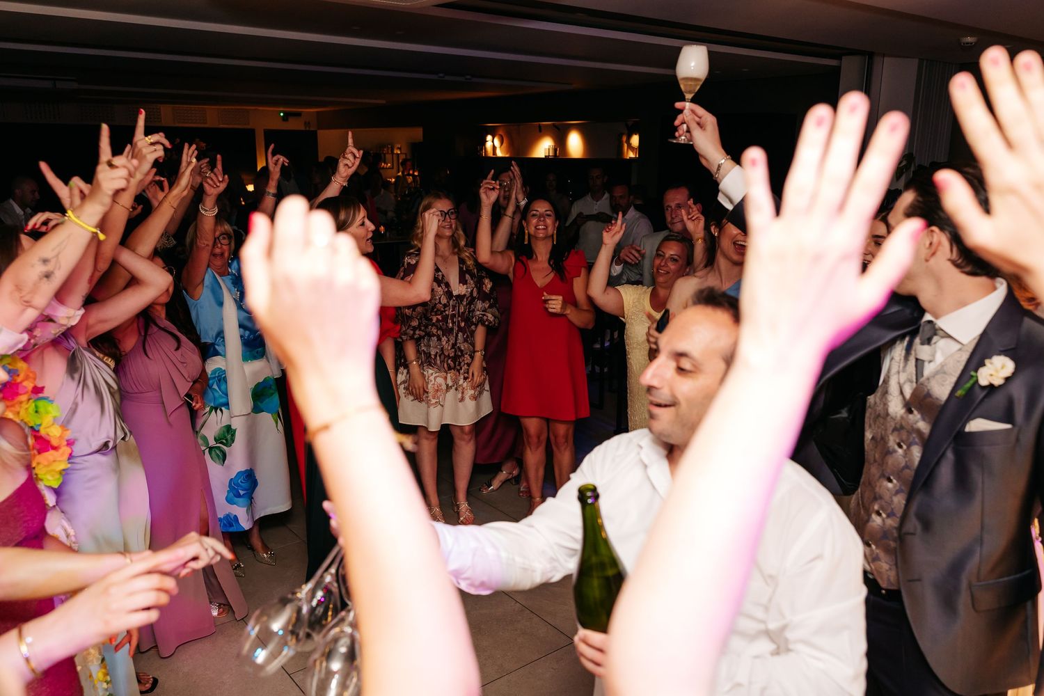 Energetic wedding reception dance floor with guests raising hands and celebrating with drinks.