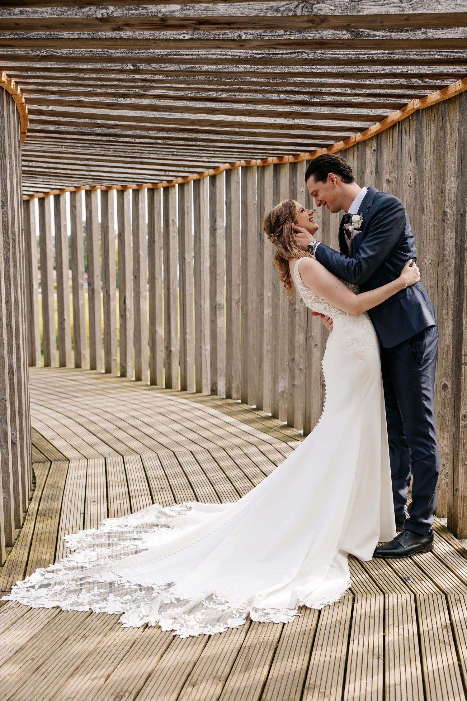 Bride and groom share a romantic moment in a wooden corridor, her lace gown trailing elegantly on the deck.
