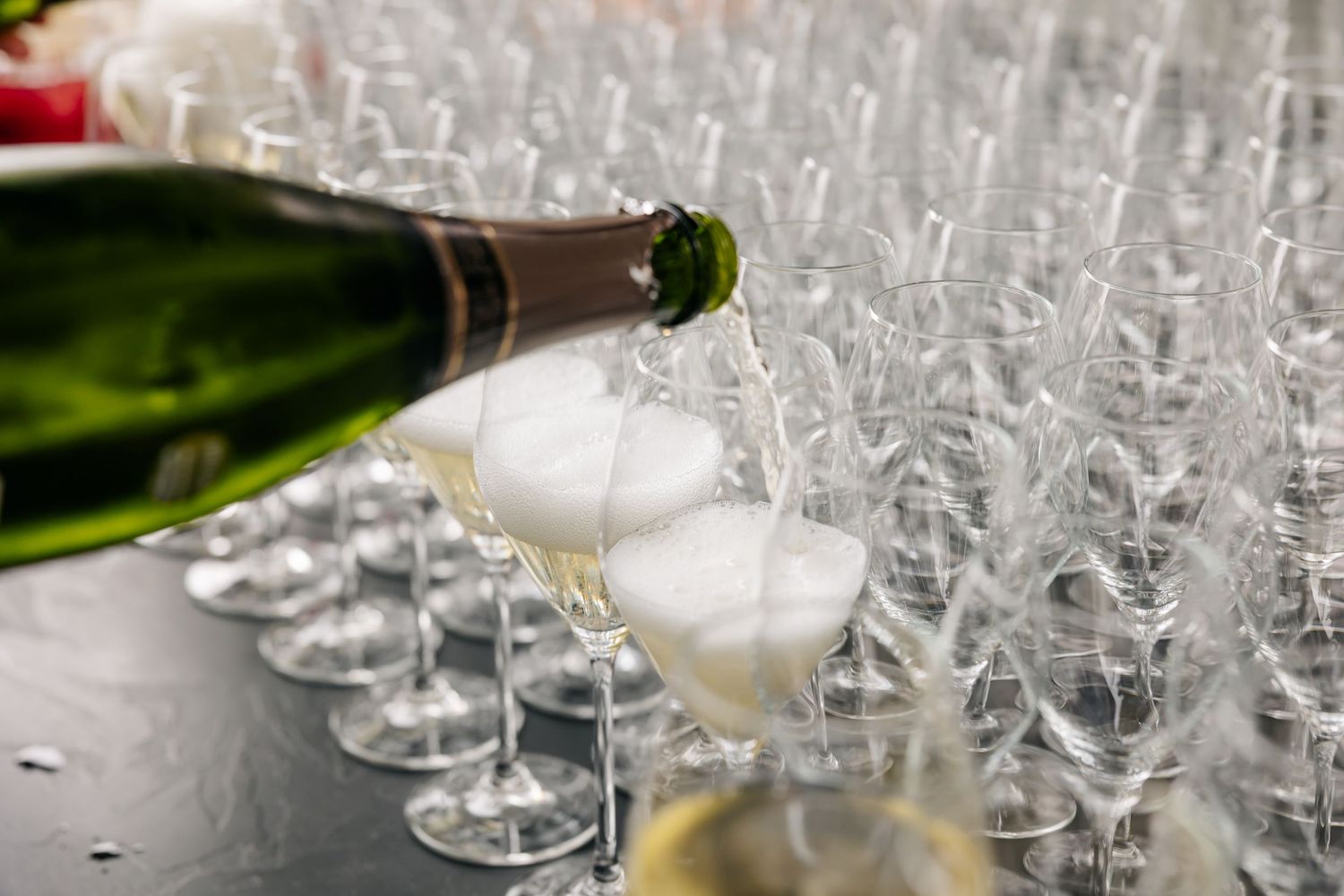 Champagne being poured into a coupe glass surrounded by rows of empty crystal glasses at a wedding reception.