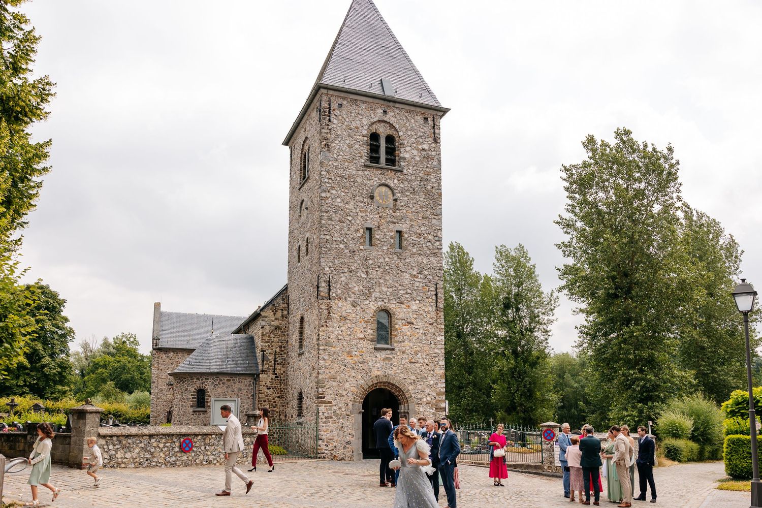 Wedding guests gather outside a historic stone church tower on a sunny day.