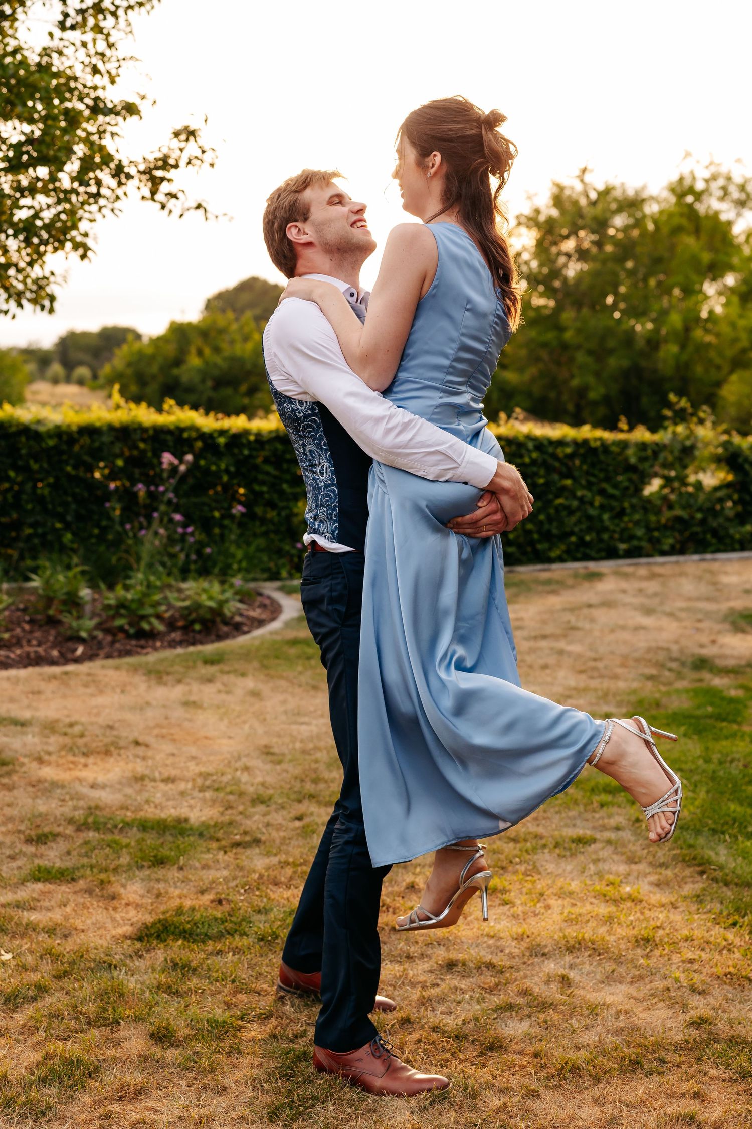 Groom lifts bride in a blue dress as they share a romantic moment in a sunlit garden at sunset.