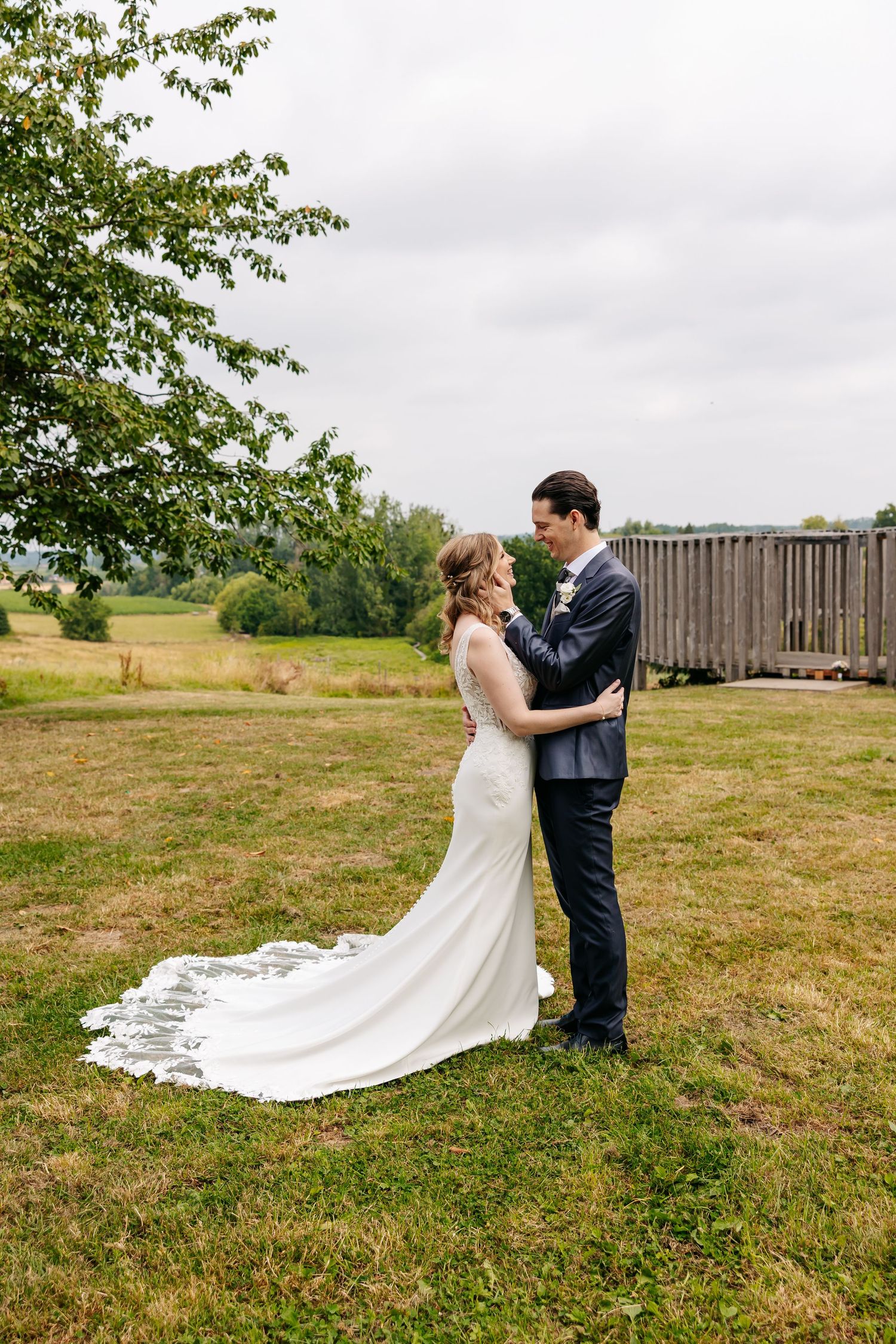 Bride and groom sharing a romantic embrace in a wide open green field with a rustic fence backdrop.