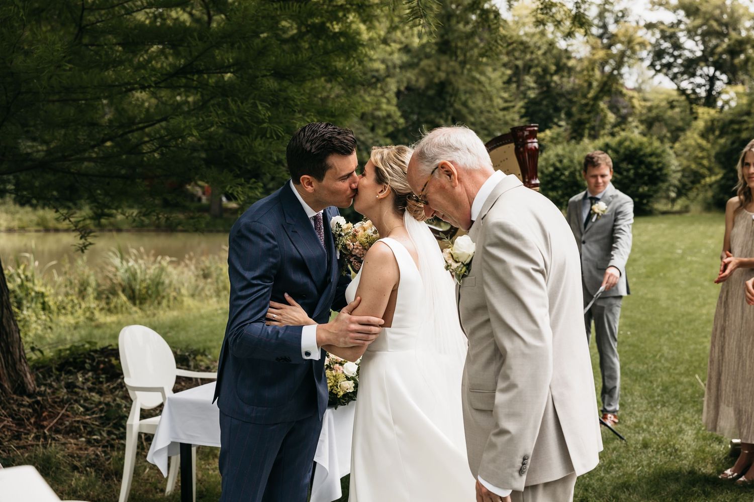 Groom in navy suit kisses bride in white gown as older gentleman congratulates them at ceremony.