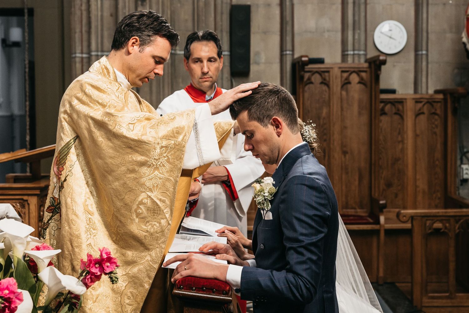 Priest in golden vestments blesses groom during Catholic wedding ceremony at decorated church altar.