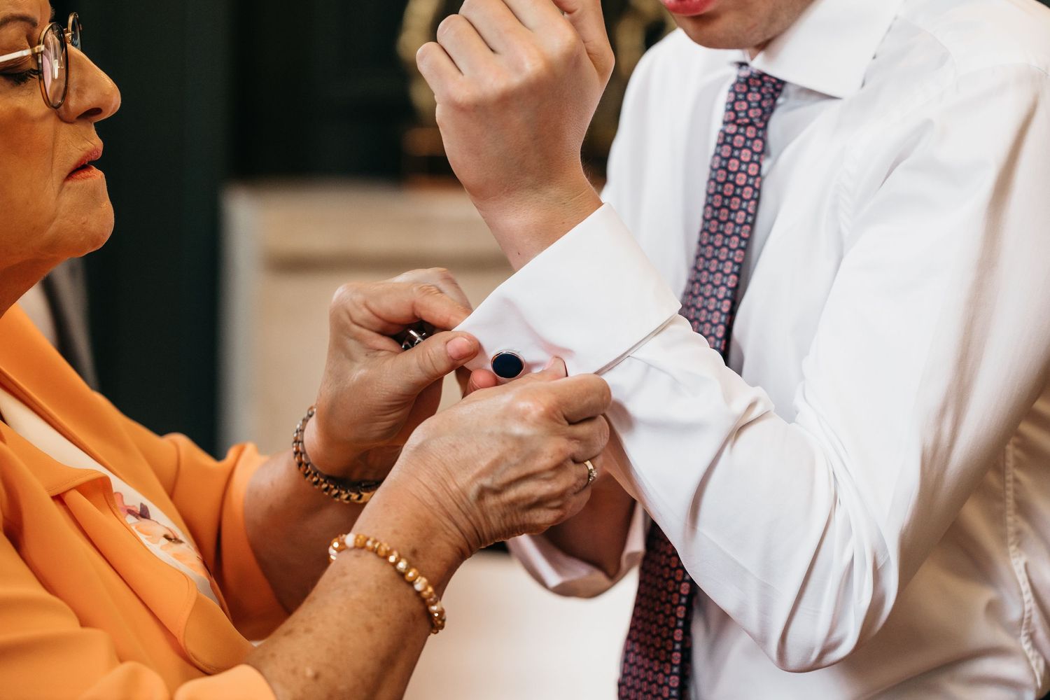 Mother fastening silver cufflinks on groom's white dress shirt sleeve before wedding ceremony.