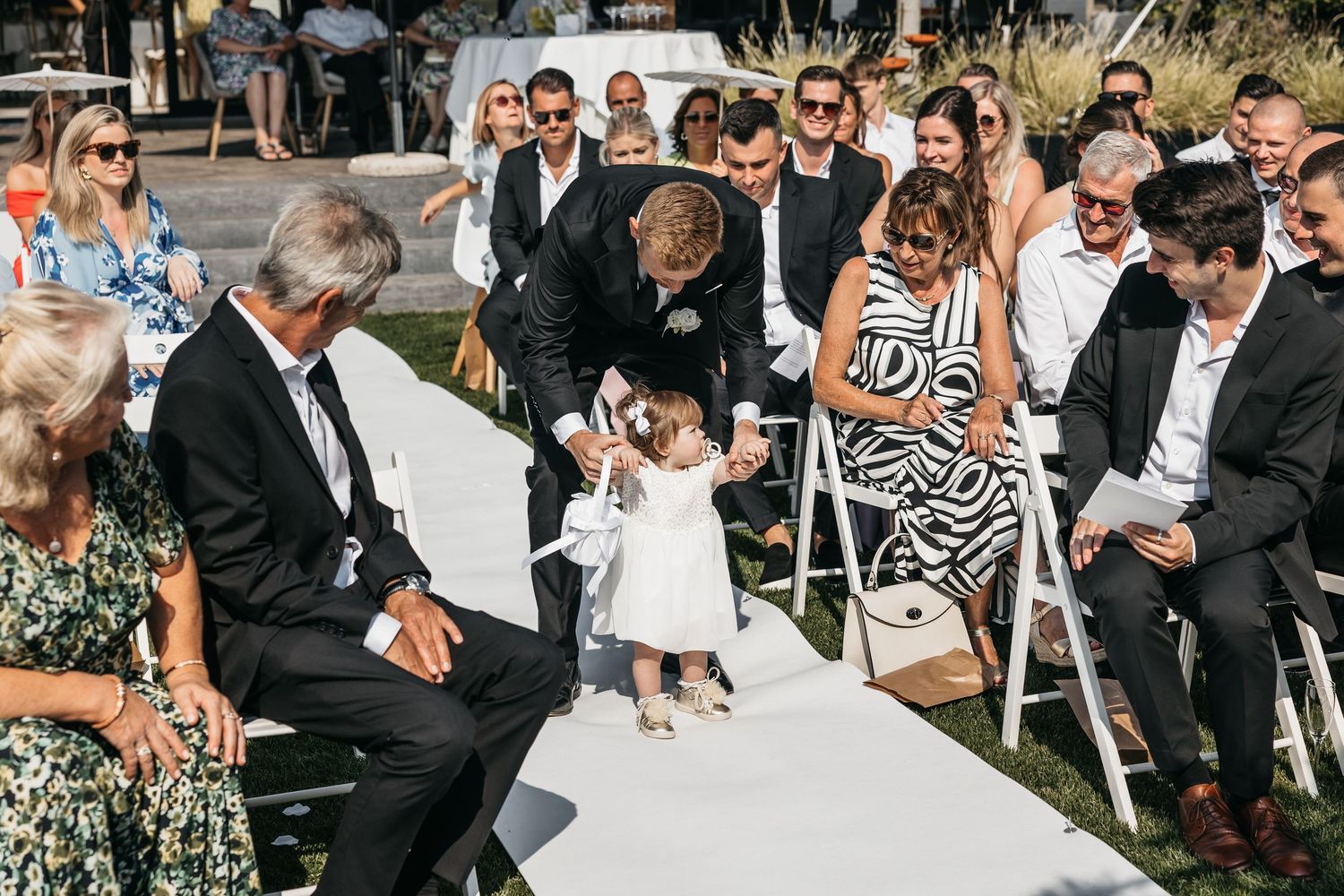 Sweet moment as groomsman guides tiny flower girl in white dress along the wedding aisle.
