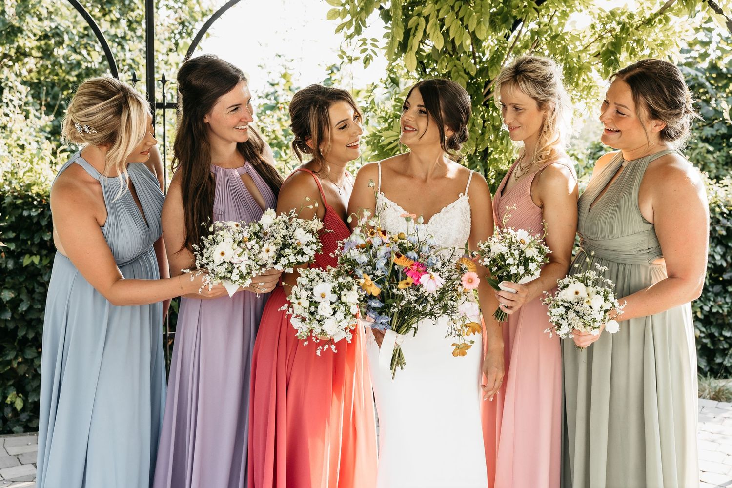 Bride and five bridesmaids in pastel mismatched gowns laughing and holding bouquets under a green floral arch.