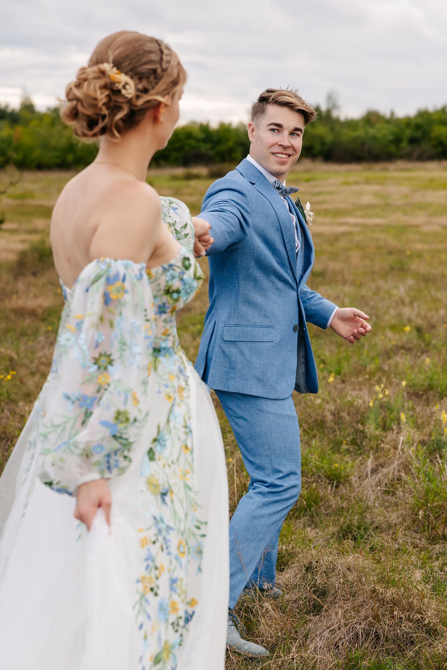 Groom in blue suit smiling at bride in floral off-shoulder wedding dress in an open countryside field.