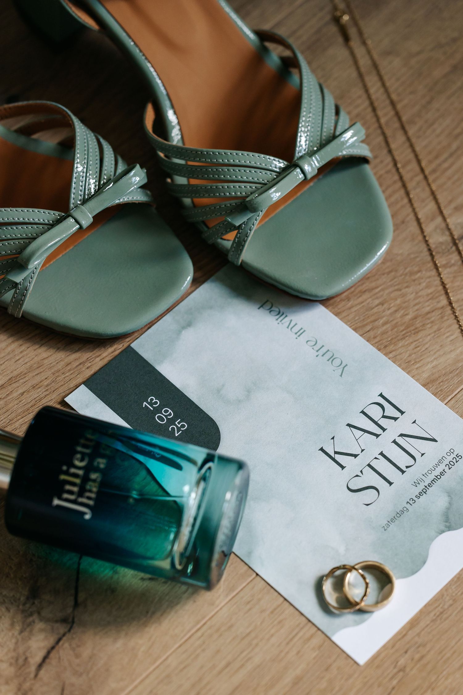 Wedding flat lay with sage green heels, perfume bottle, gold rings, and elegant invitation card.