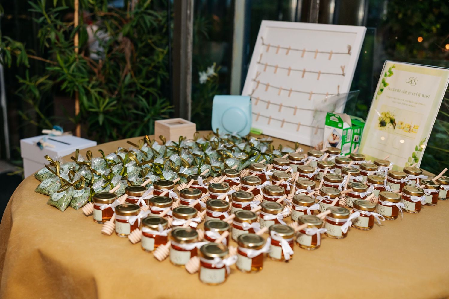 Wedding favor table displaying rows of small honey jars with greenery and a seating chart board.