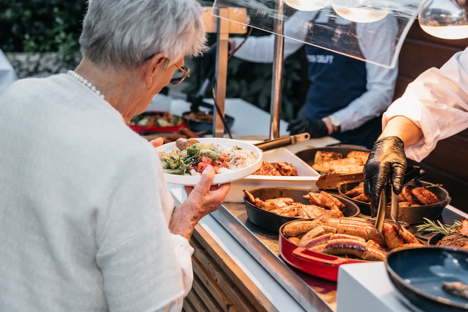 Elderly woman being served grilled sausages and meats at an outdoor catering buffet station.
