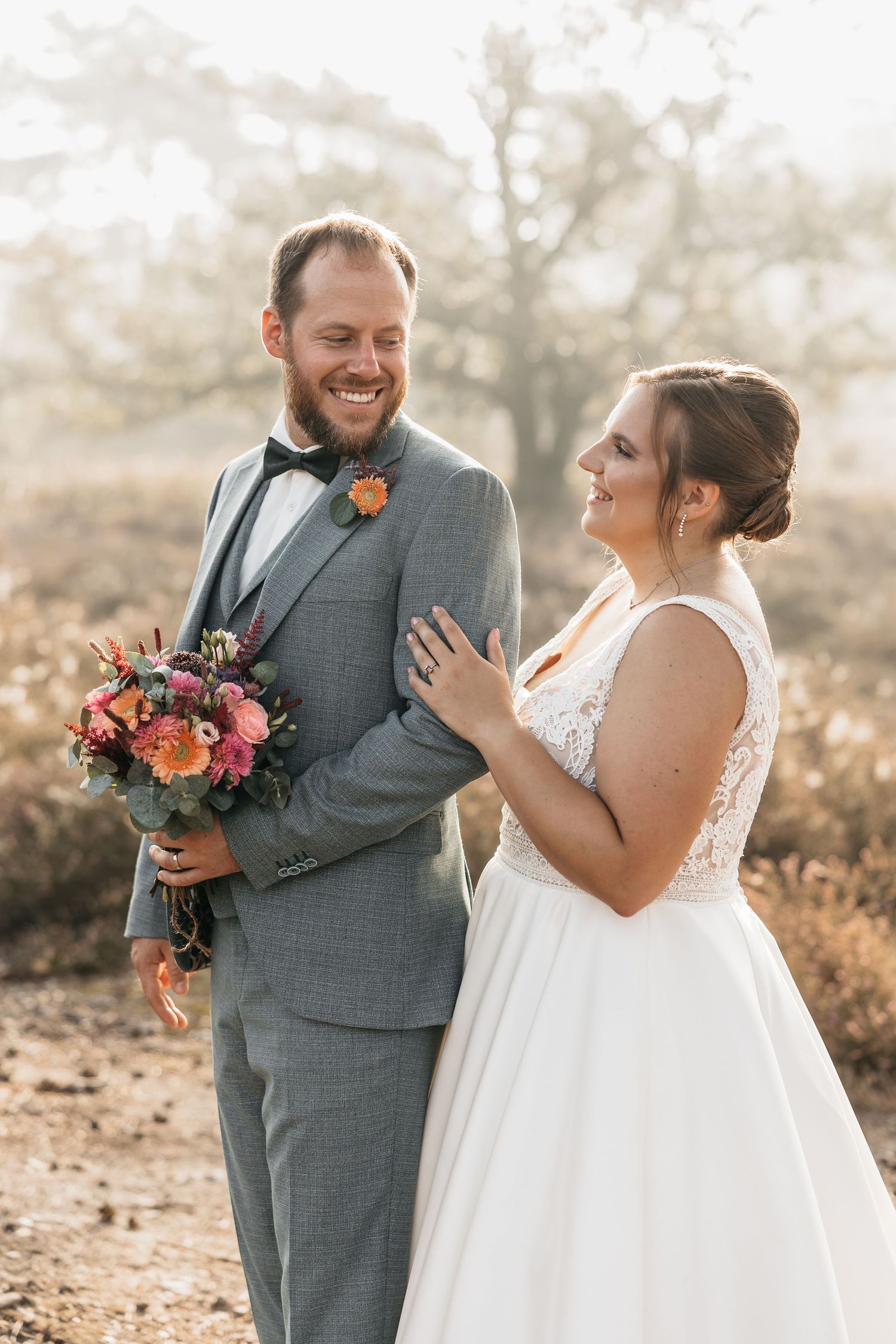 Laughing bride and groom share a joyful moment outdoors, groom holding vibrant bridal bouquet in warm sunlight.