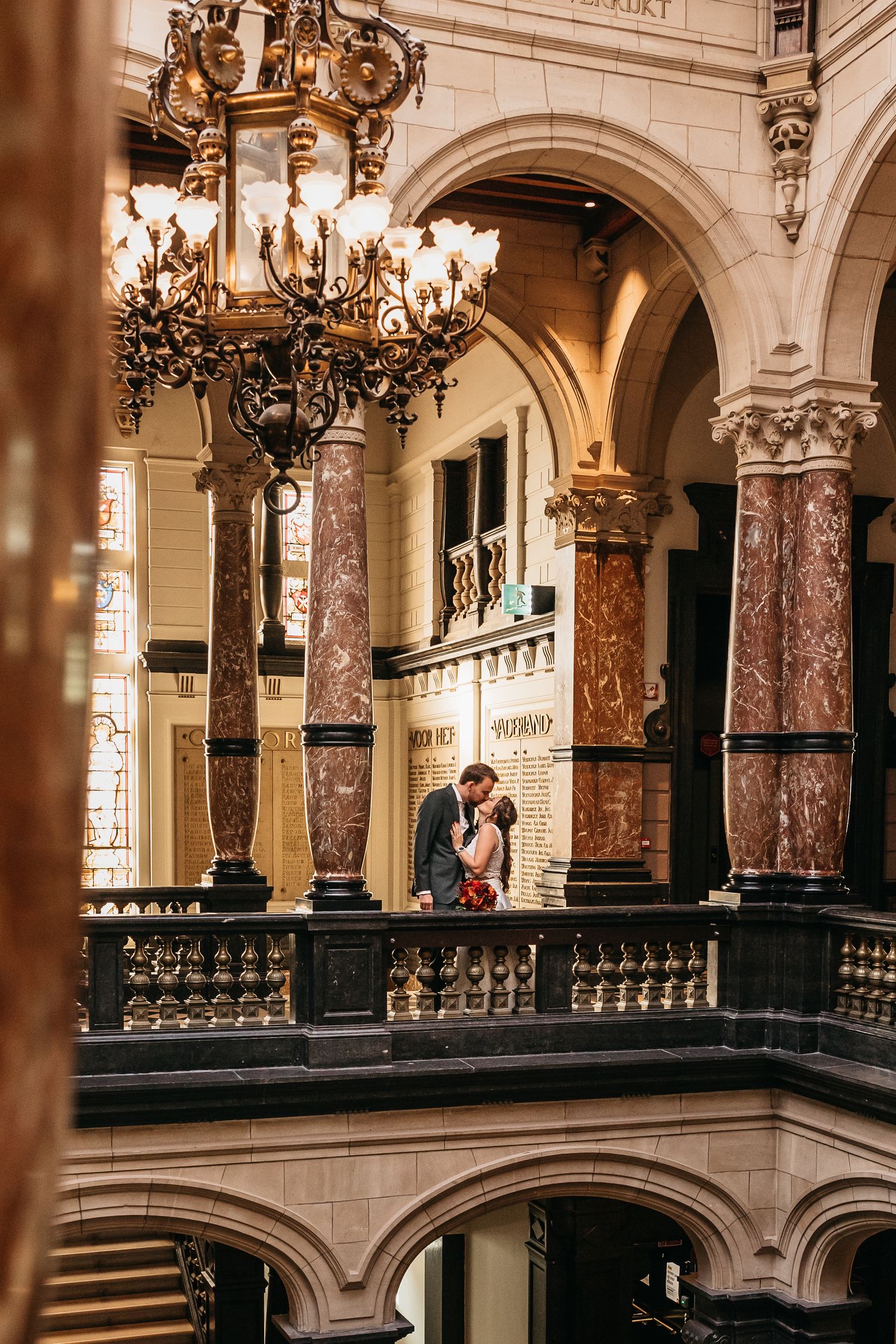 Couple kissing on balcony surrounded by grand chandeliers and marble columns.