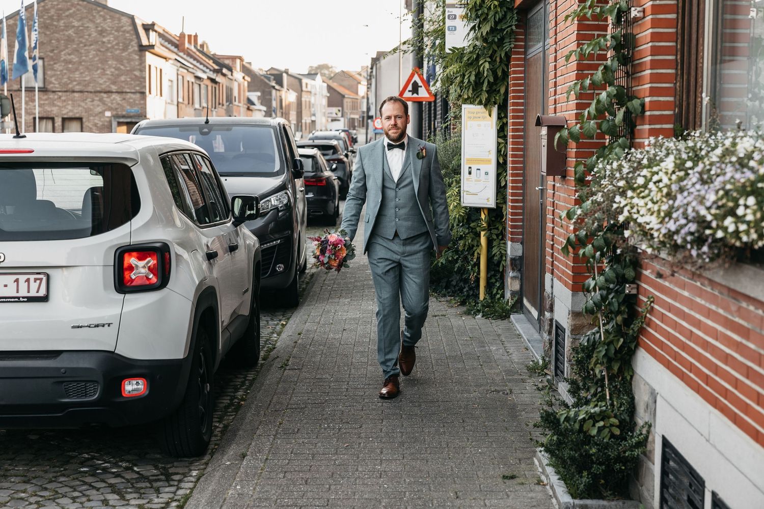 Groom in grey three-piece suit walking along a cobblestone street beside parked cars.