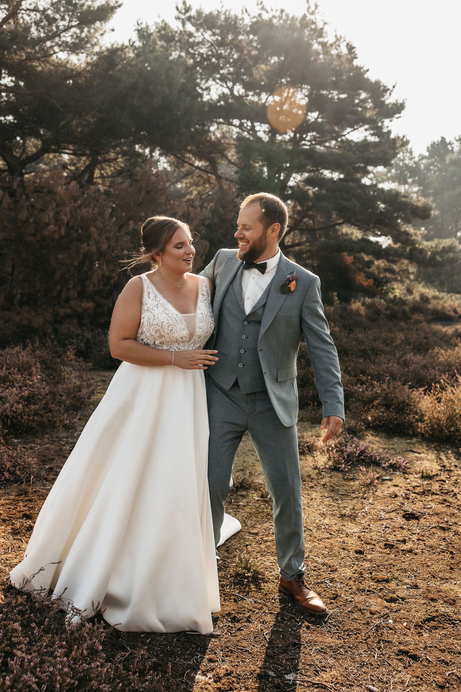 Smiling newlyweds walk together through golden heathland landscape with pine trees glowing in warm sunlight.