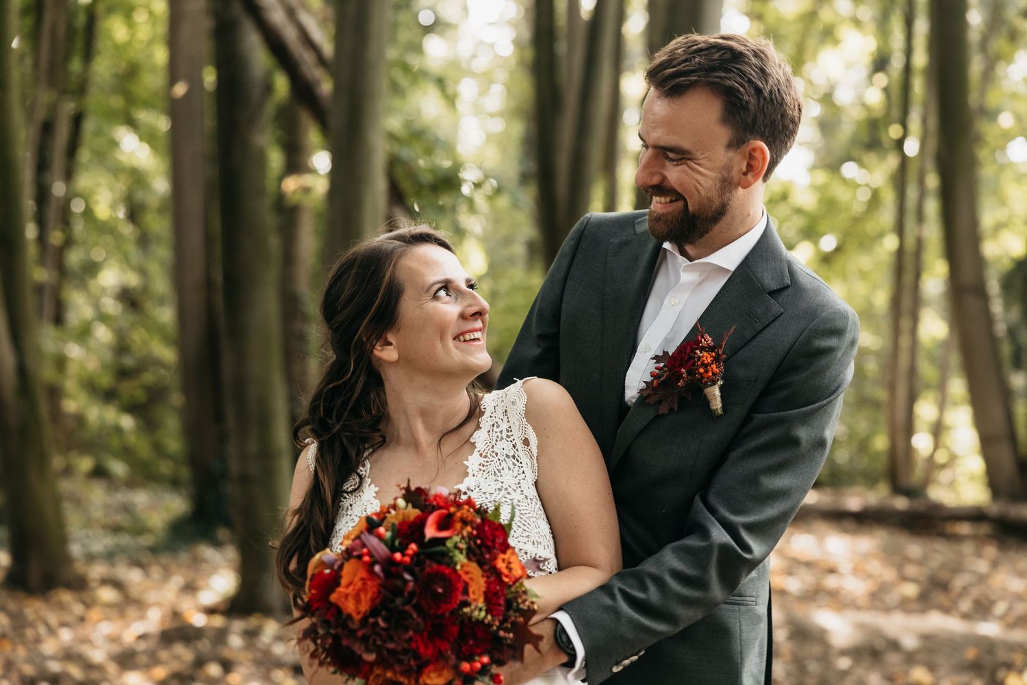 Happy bride and groom sharing a joyful moment with red bouquet in autumn woodland.