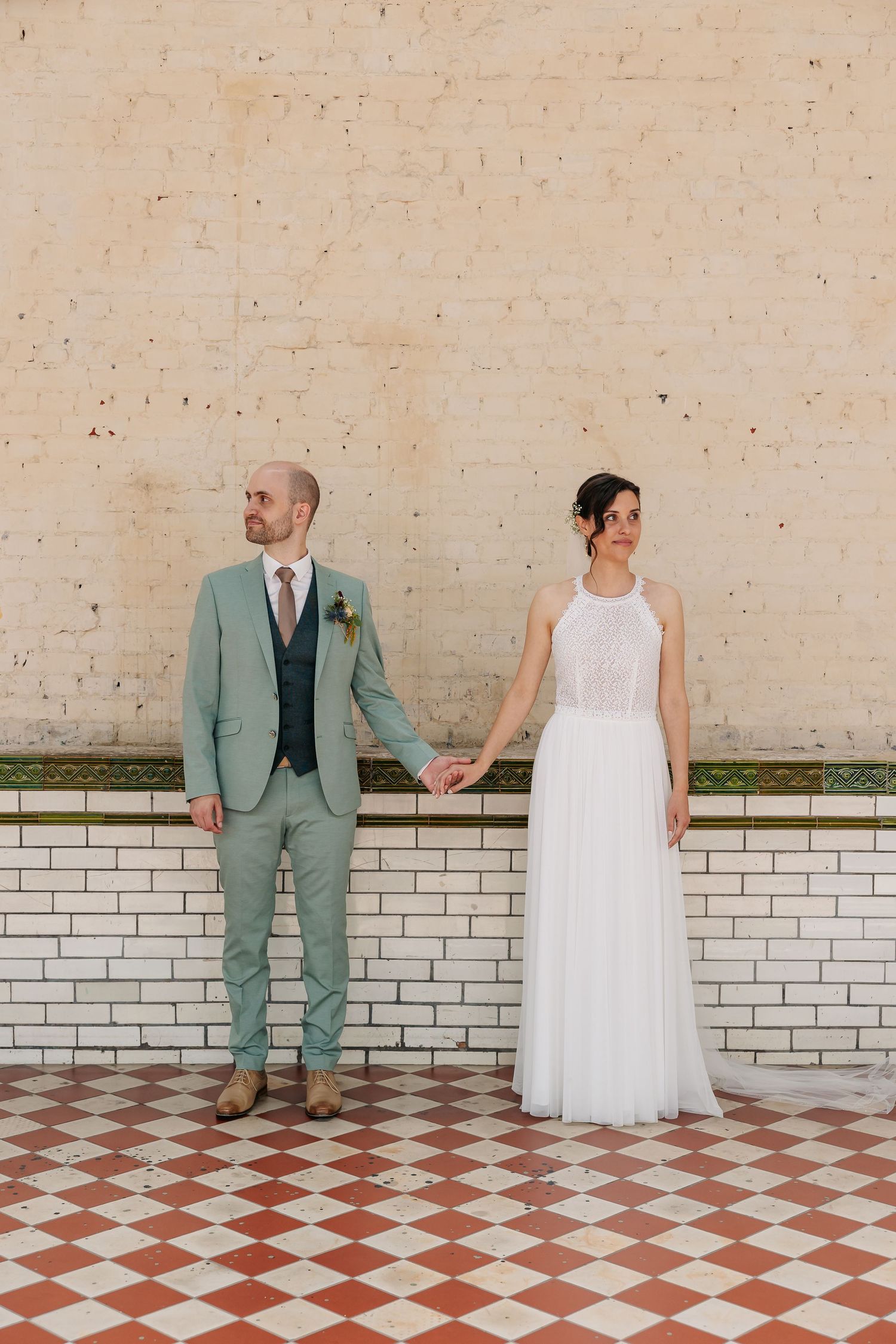 Bride and groom hold hands standing apart before a weathered brick wall on checkered tile floor.