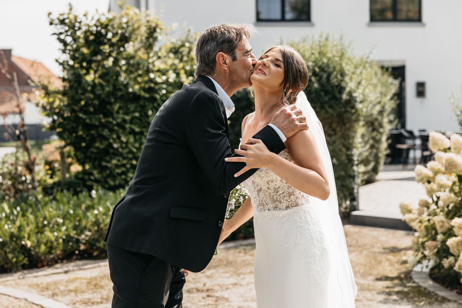 Groom joyfully lifting bride in white lace wedding dress during outdoor wedding celebration in garden setting.