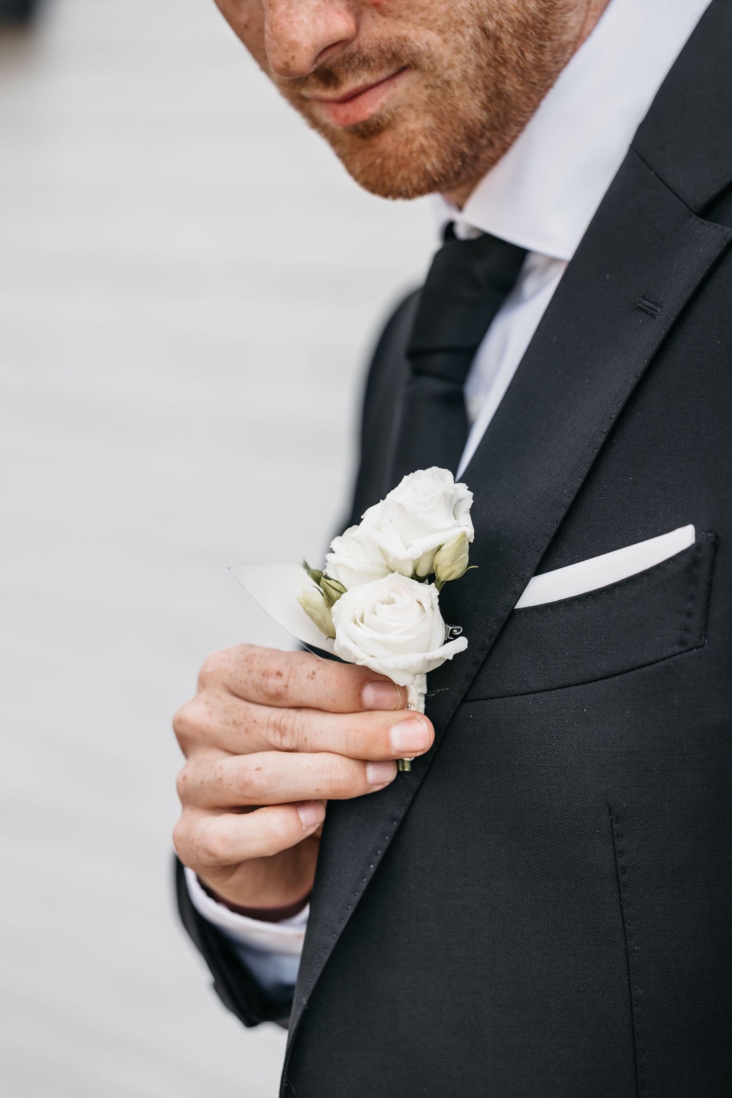 Groom in black suit with black tie adjusting white rose boutonniere with white pocket square.