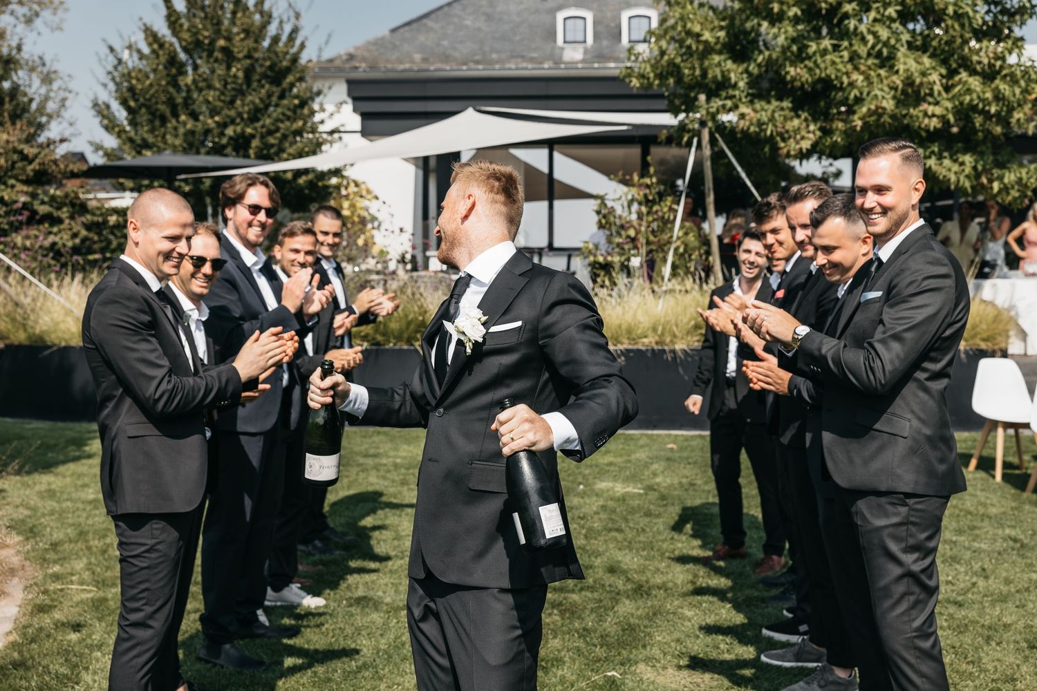 Groom holding champagne bottle while groomsmen in black suits clap and celebrate outdoors.