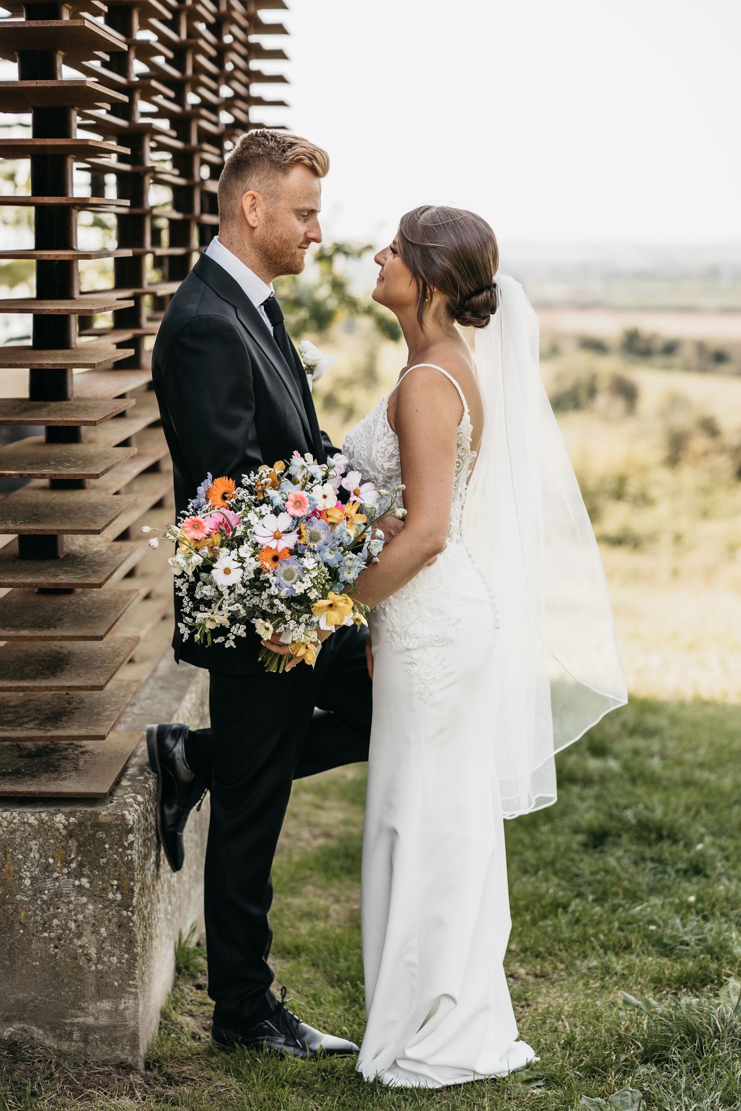Bride and groom share an intimate moment by a wooden structure with colorful bridal bouquet.