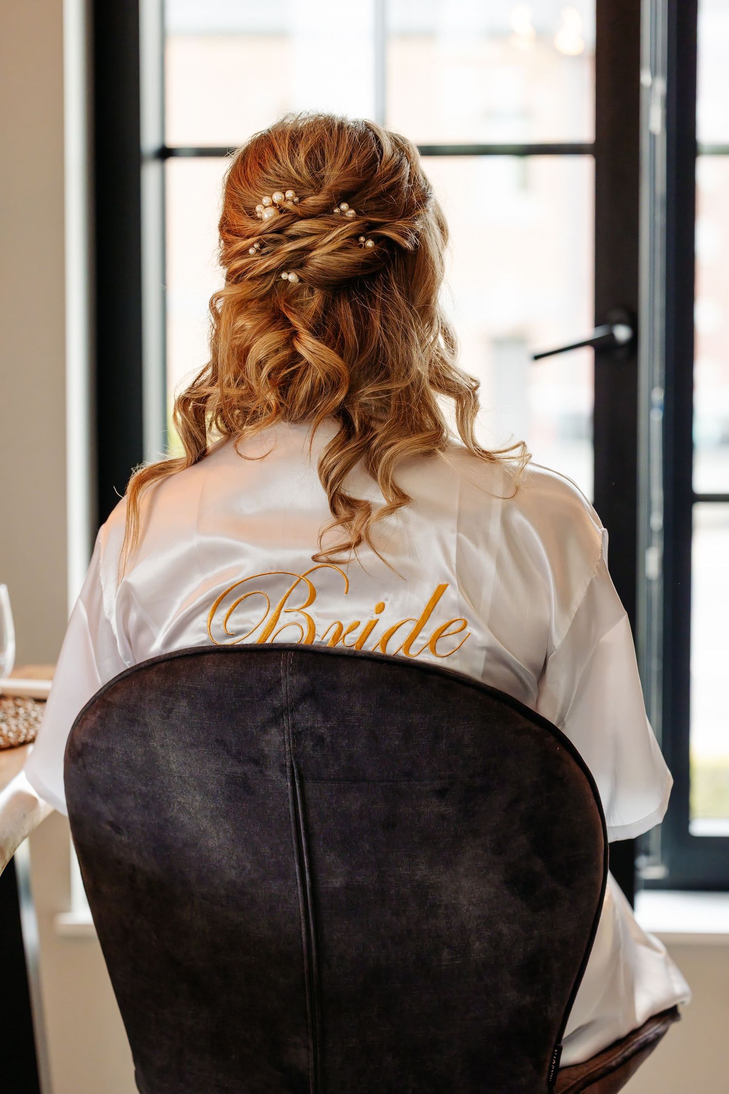 Back view of bride in white satin robe showcasing elegant half-up hairstyle with floral hair pins.
