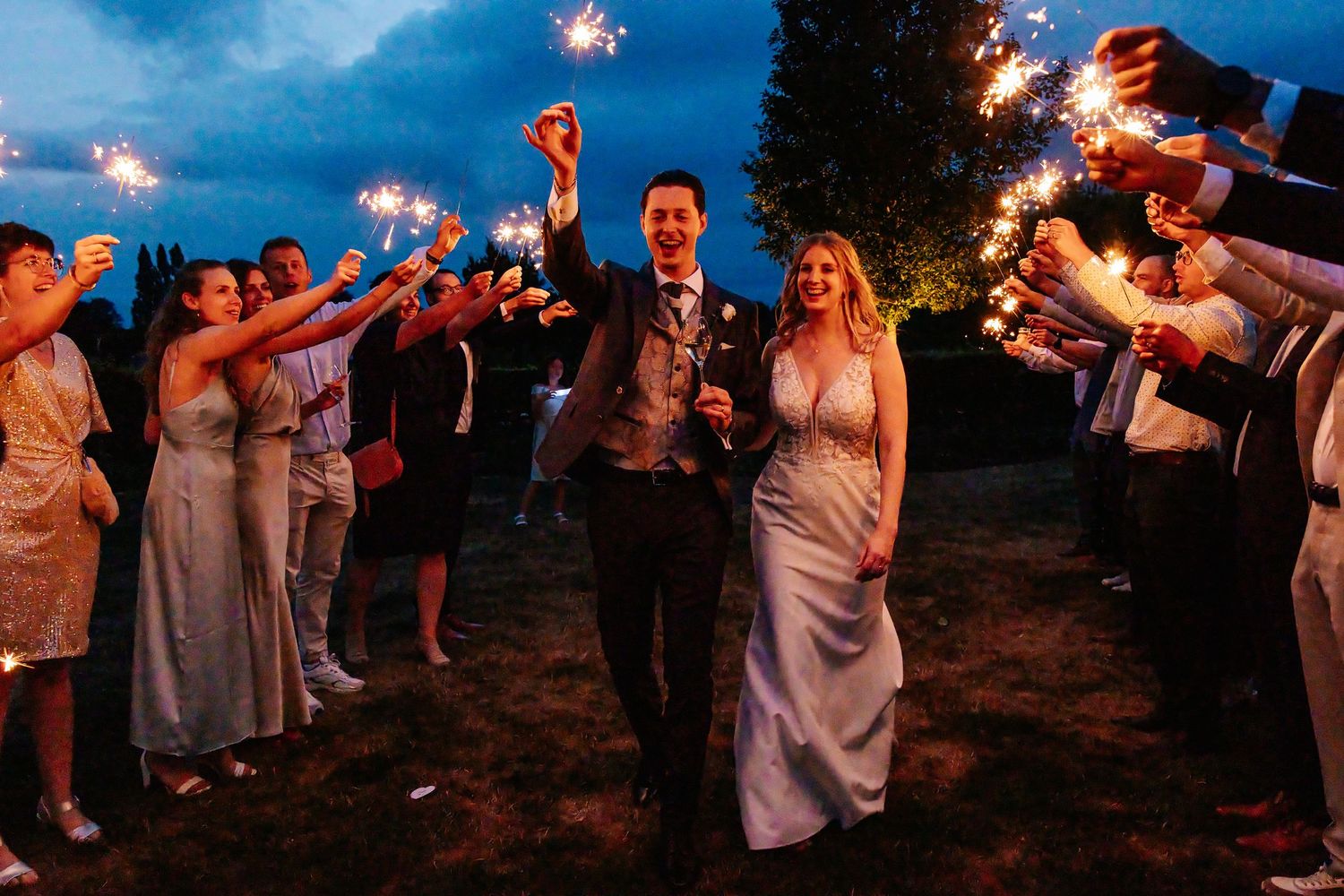 Newlyweds walk through sparkler tunnel with guests at twilight wedding send-off celebration.