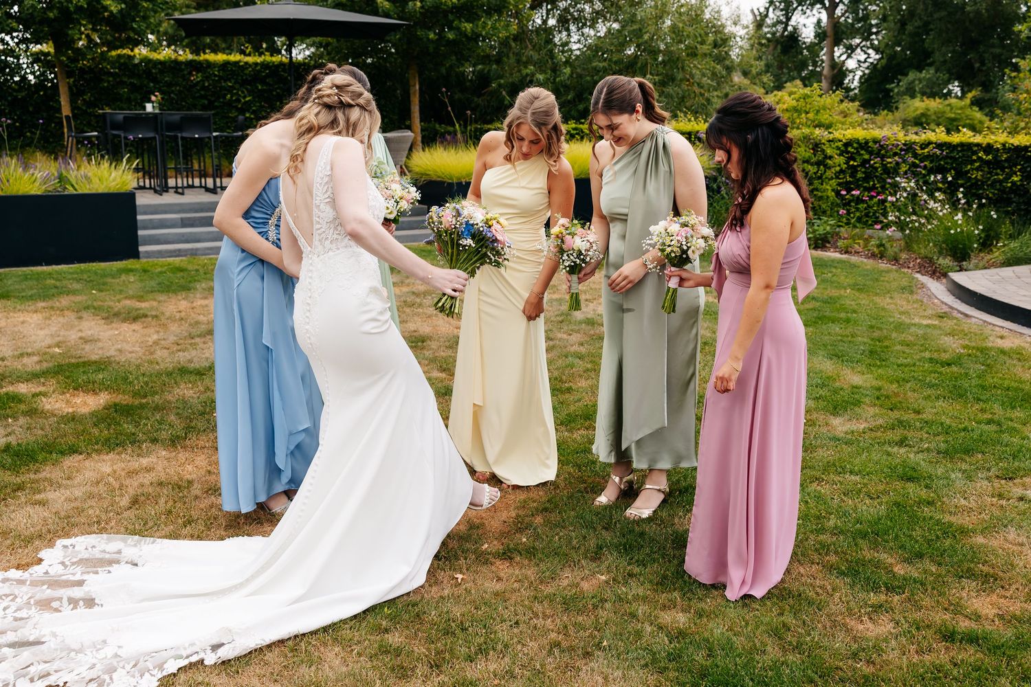 Bridesmaids in pastel yellow, blue, and green gowns admire bride's lace wedding dress train in garden.