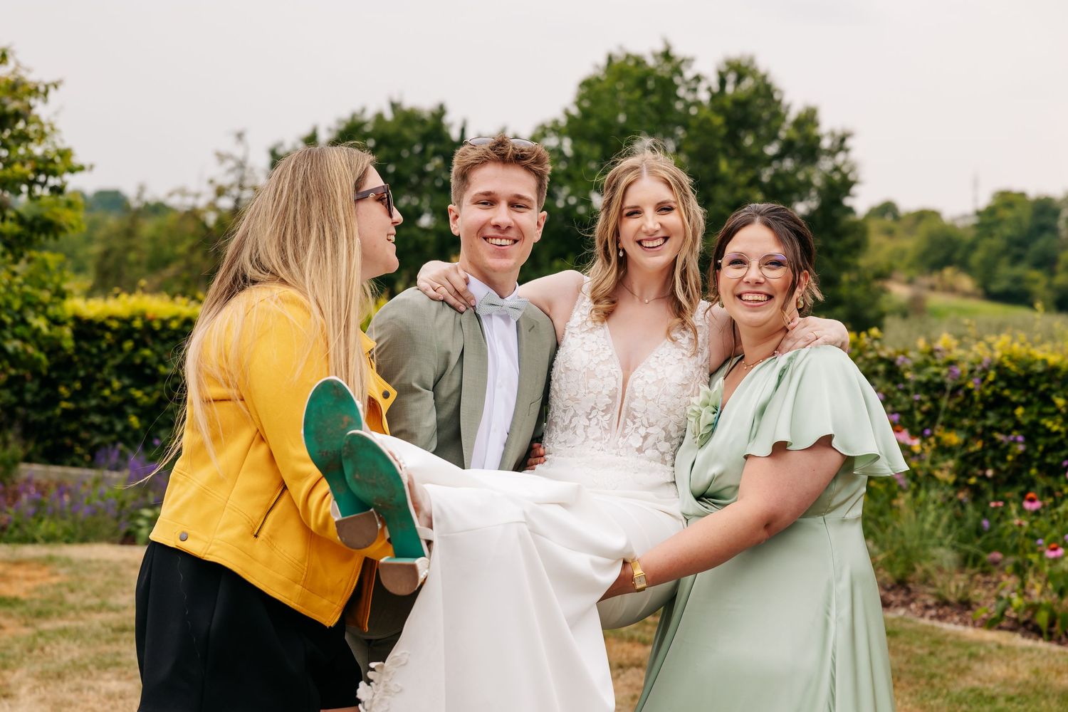 A bride in a white lace gown laughs with three friends, one holding her dress train, in a garden.