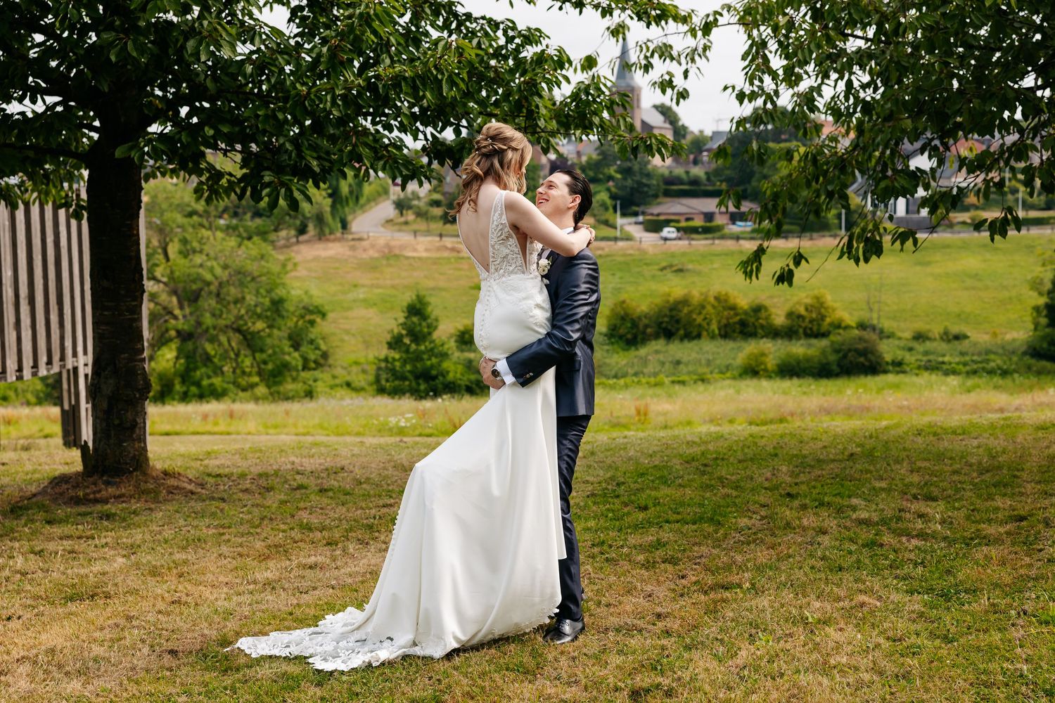 Bride and groom embracing tenderly under a tree in a scenic green outdoor wedding setting.