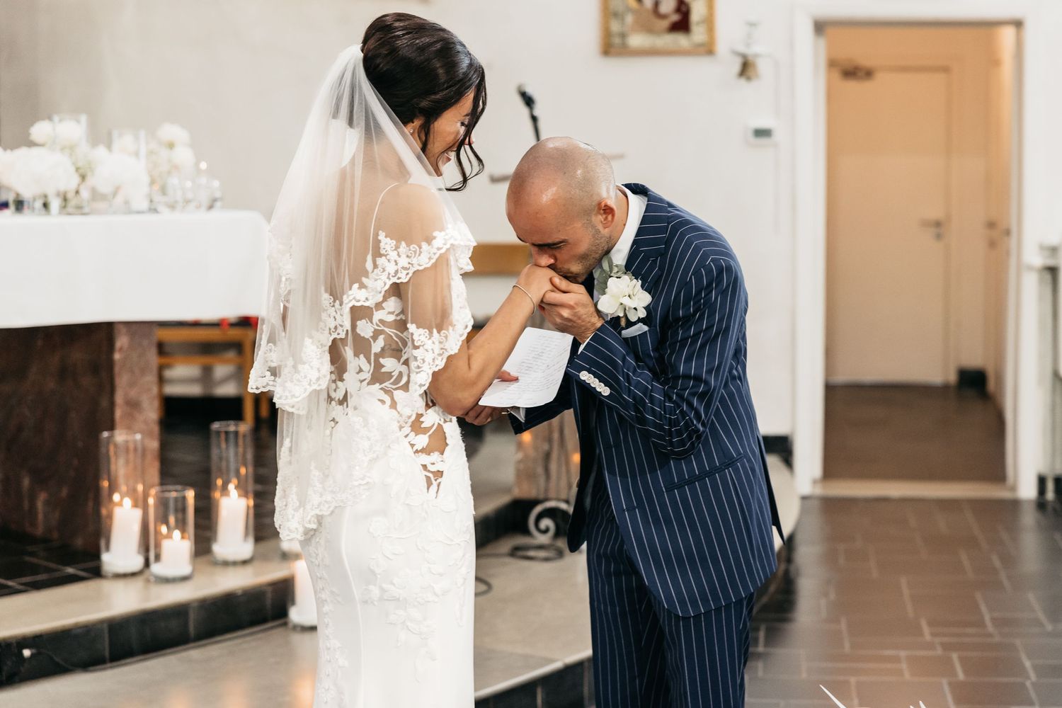 Groom in navy pinstripe suit kisses bride's hand during emotional wedding ceremony.