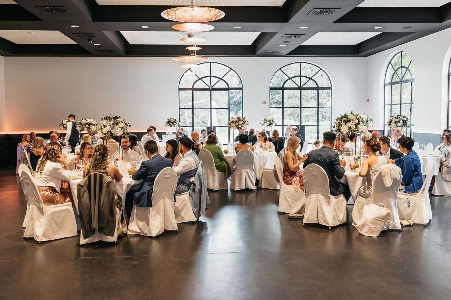 Wide view of wedding reception hall with guests seated at round tables with floral decor.