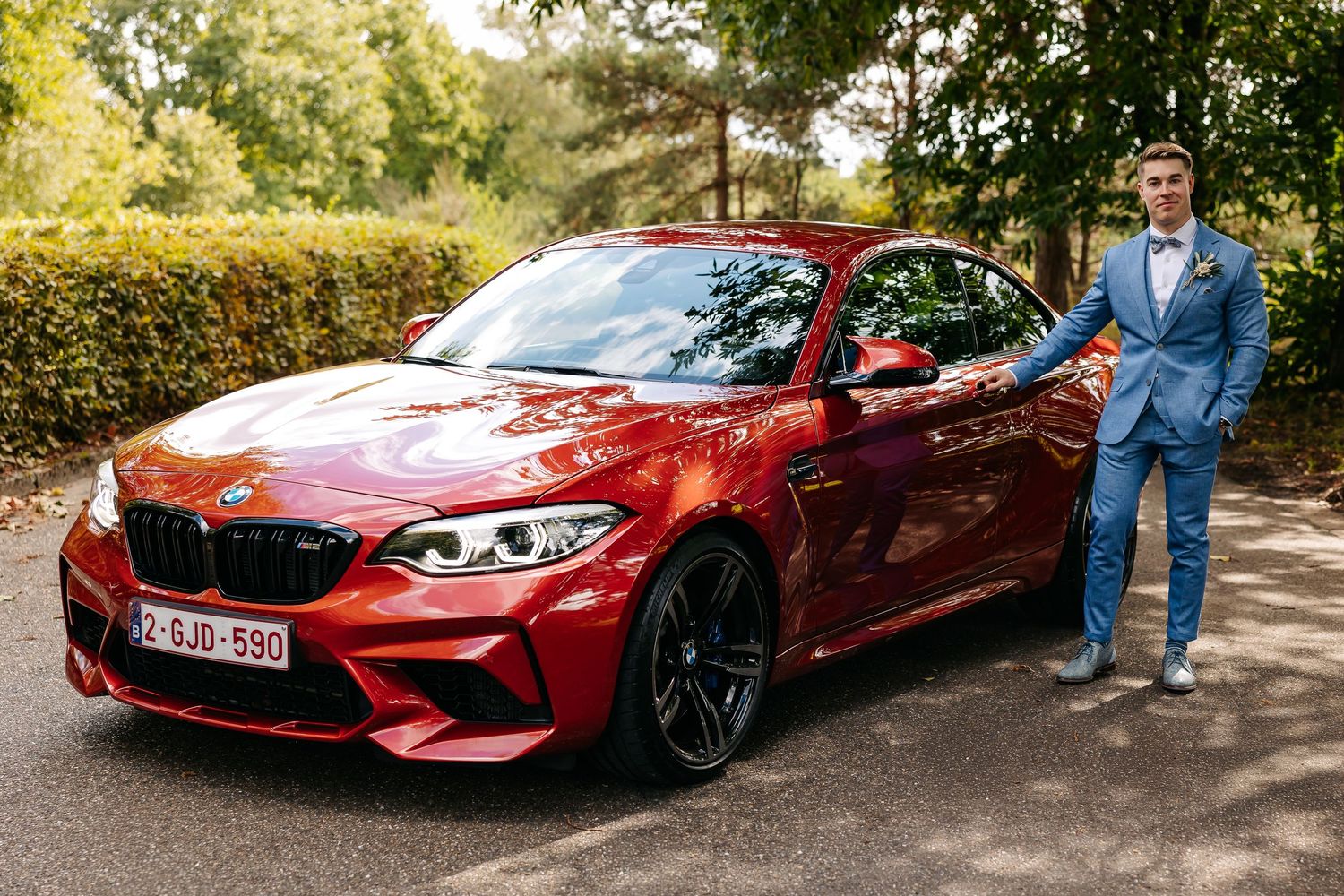 A groom in a blue suit poses proudly beside a red BMW M2 sports car on a tree-lined road.