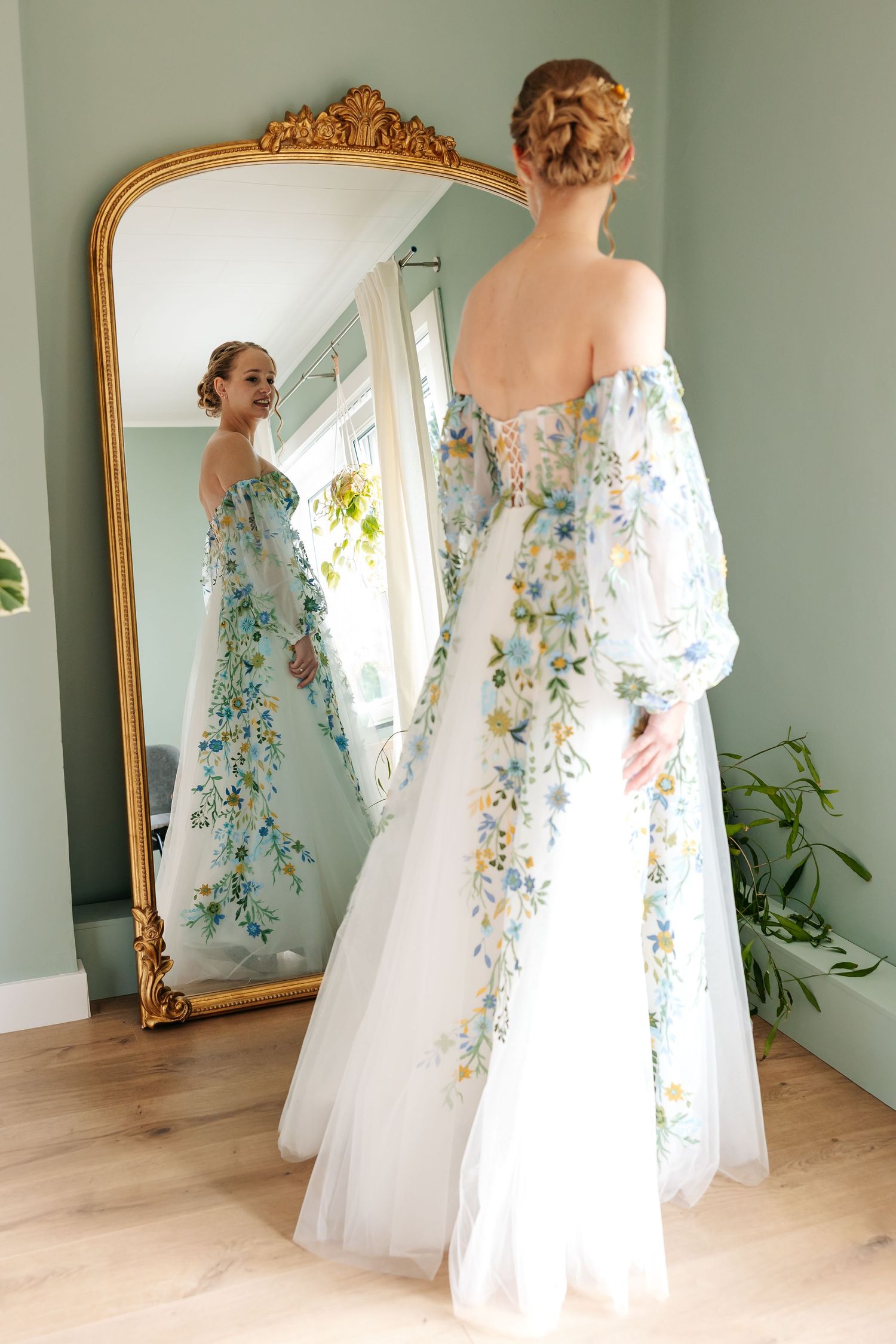 Bride in stunning floral embroidered off-shoulder wedding gown viewed from behind in gold mirror.
