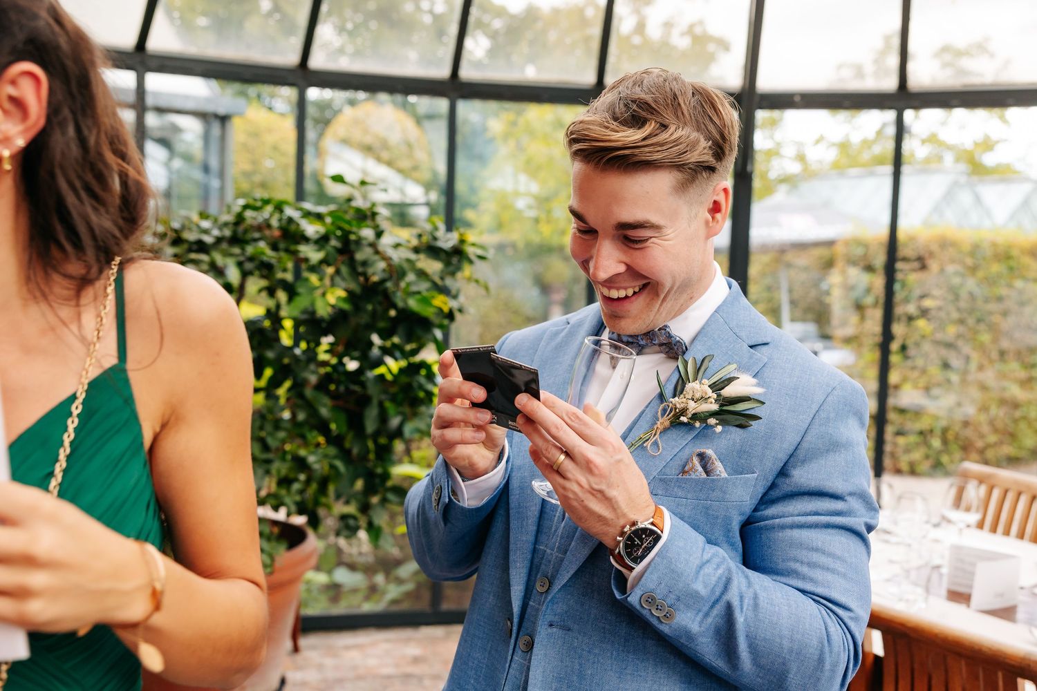 Groom in light blue suit laughing at phone screen at outdoor wedding reception.
