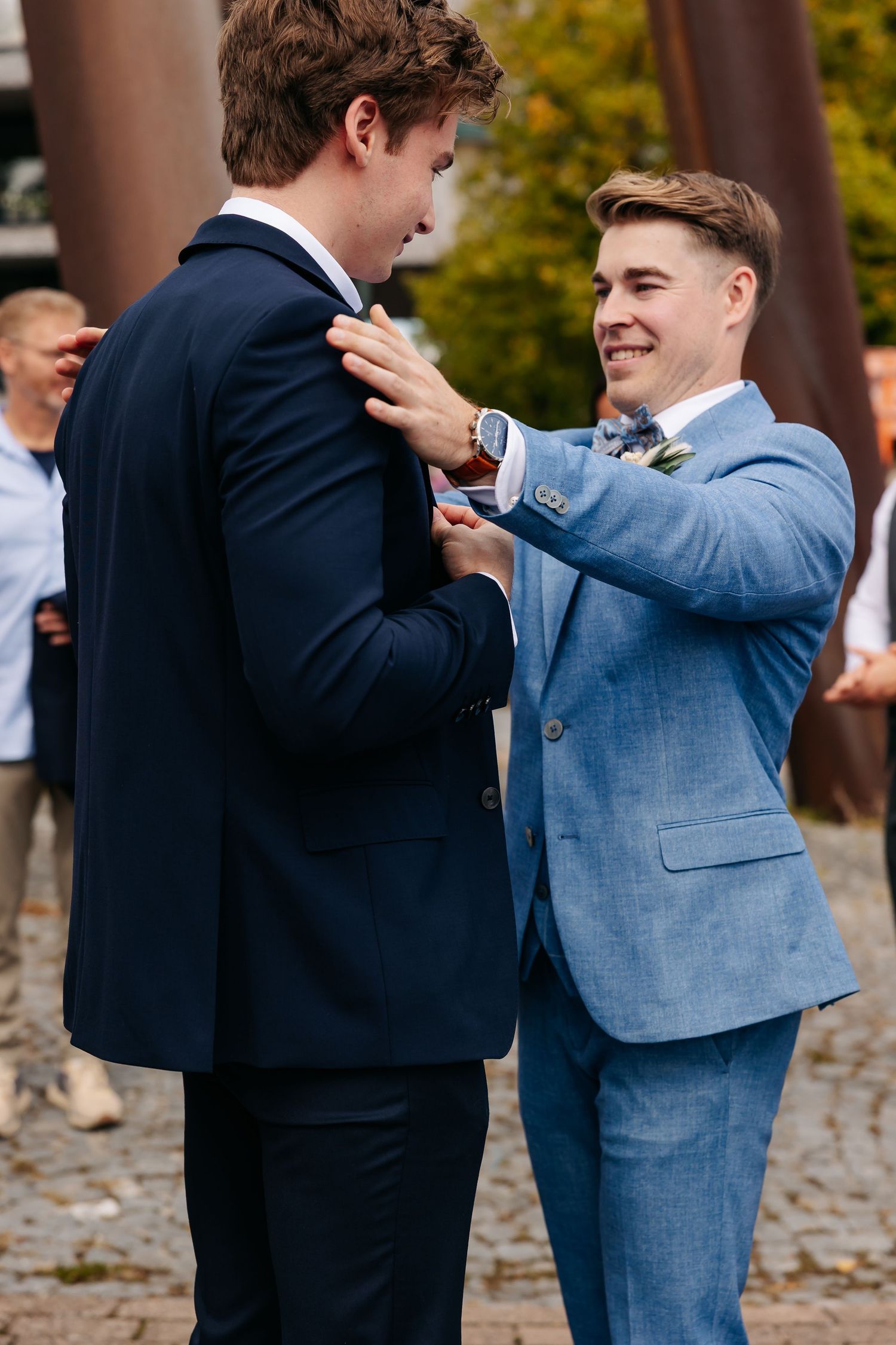 Groomsman in blue suit adjusting boutonniere on groom in navy suit at outdoor wedding.