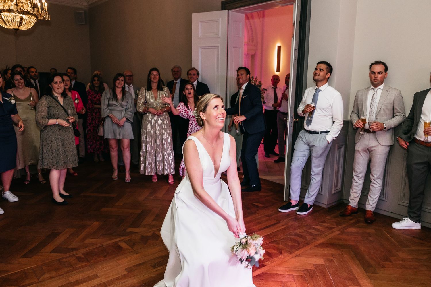 Bride in white gown prepares to toss bouquet to eager guests at wedding reception.