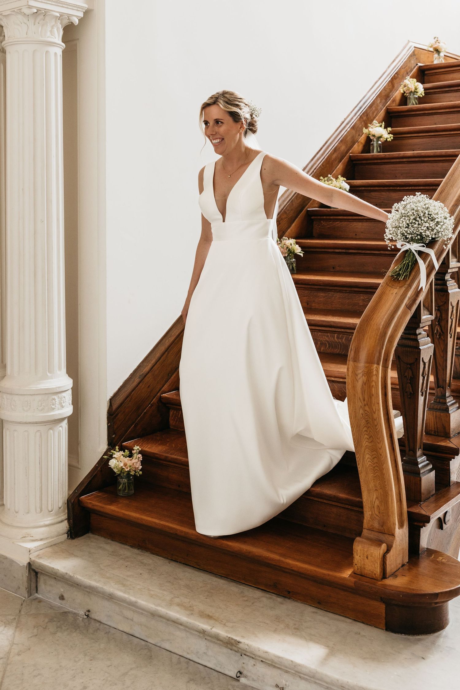 Bride in white V-neck gown descending flower-adorned wooden staircase.