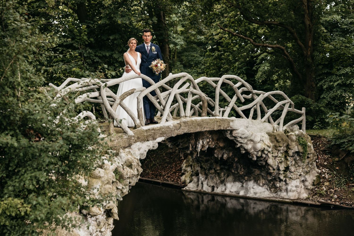 Bride and groom posing on a rustic stone bridge surrounded by lush green trees.