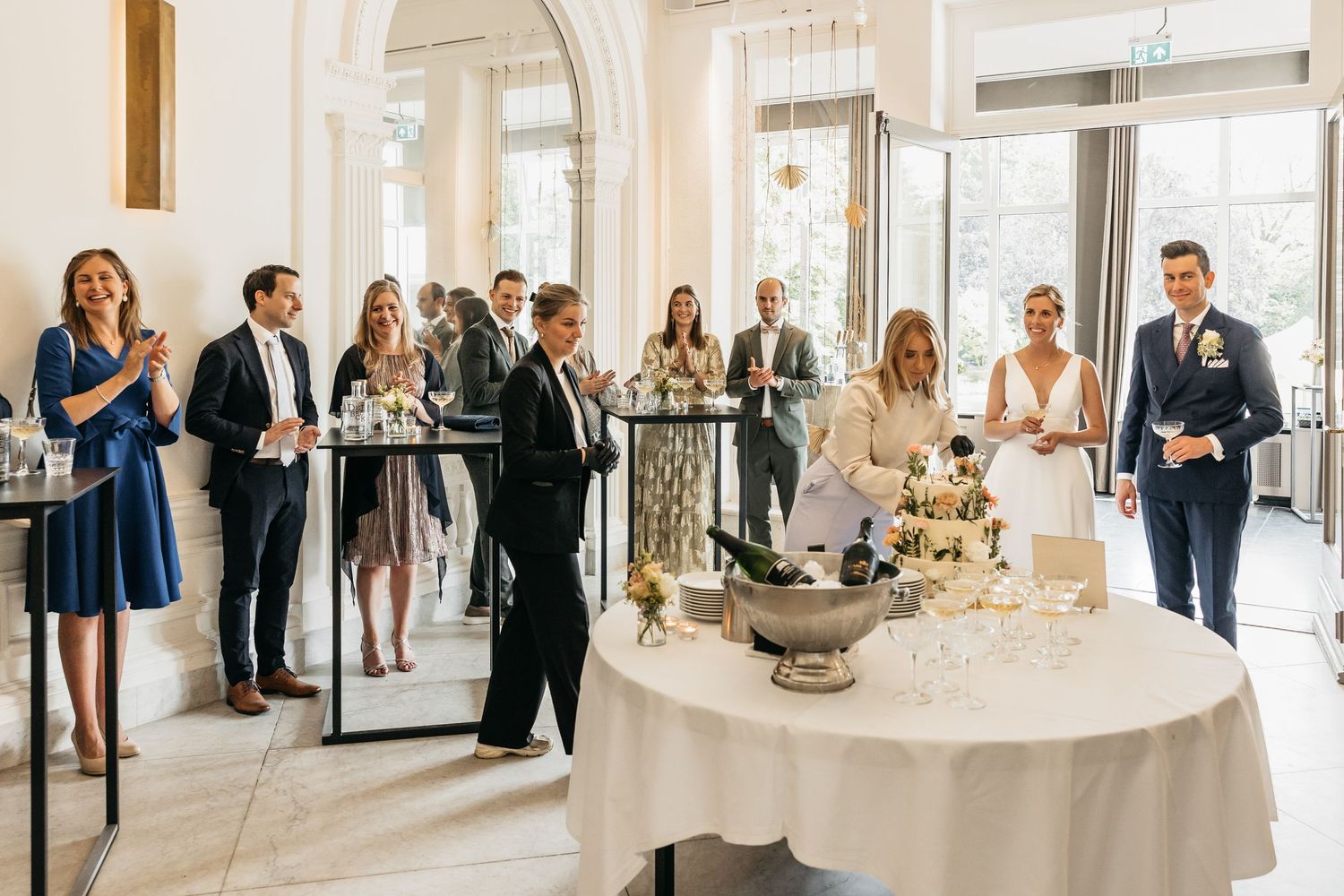 Wedding guests applauding as bride and groom stand beside champagne tower and floral arrangements.