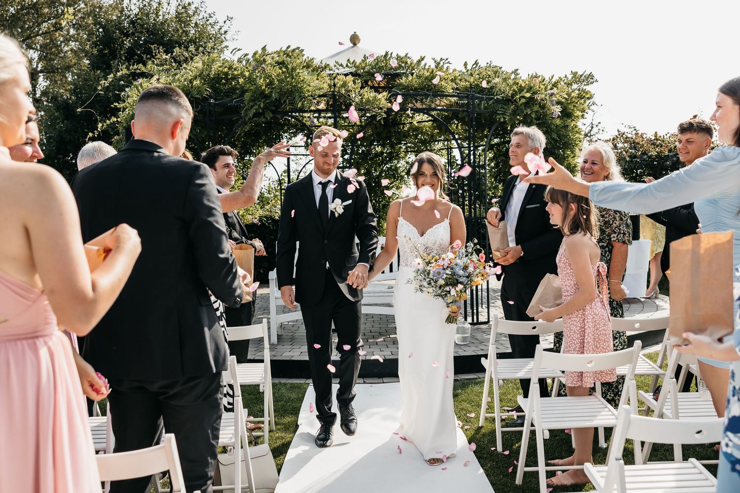Joyful newlyweds walk down aisle as guests celebrate with confetti at outdoor wedding.