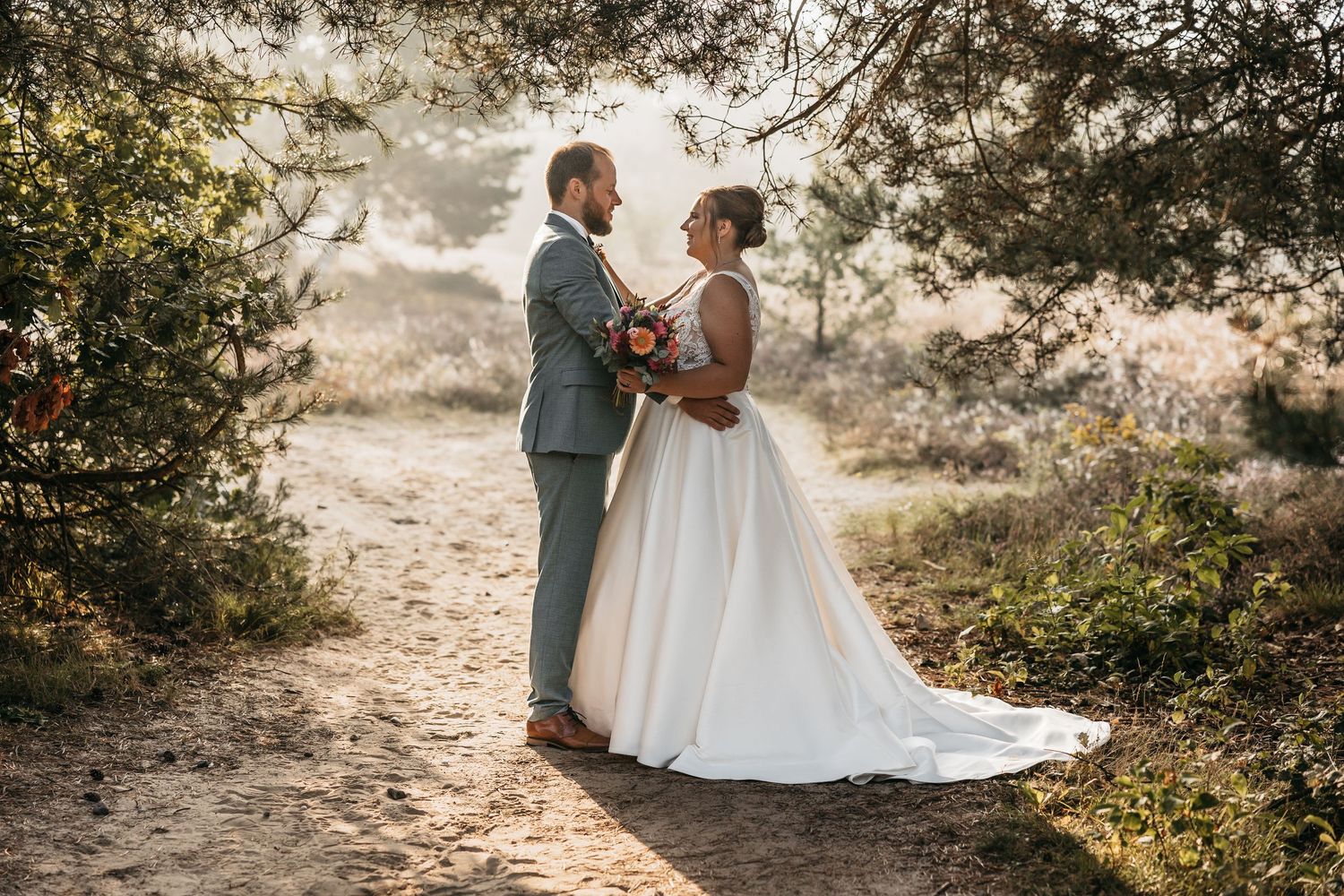 Bride and groom stand face to face on a sunlit forest trail.
