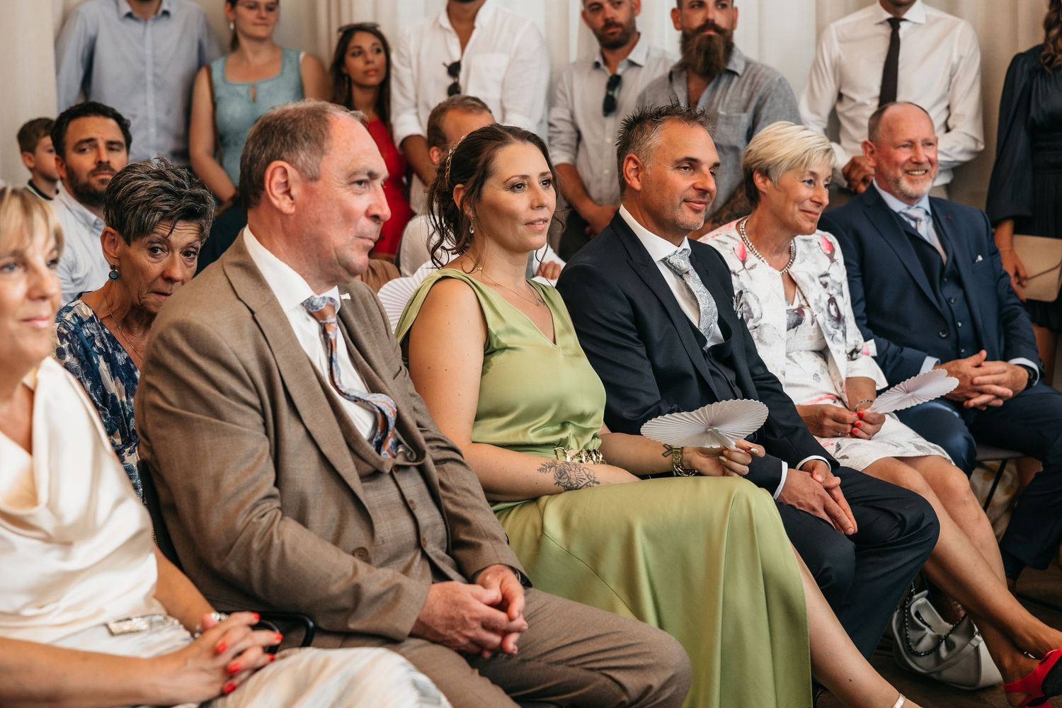 Wedding guests in formal attire seated attentively during indoor ceremony in sunlit room.