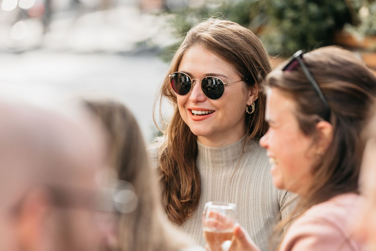 Smiling young woman wearing sunglasses enjoying drinks at an outdoor social gathering.