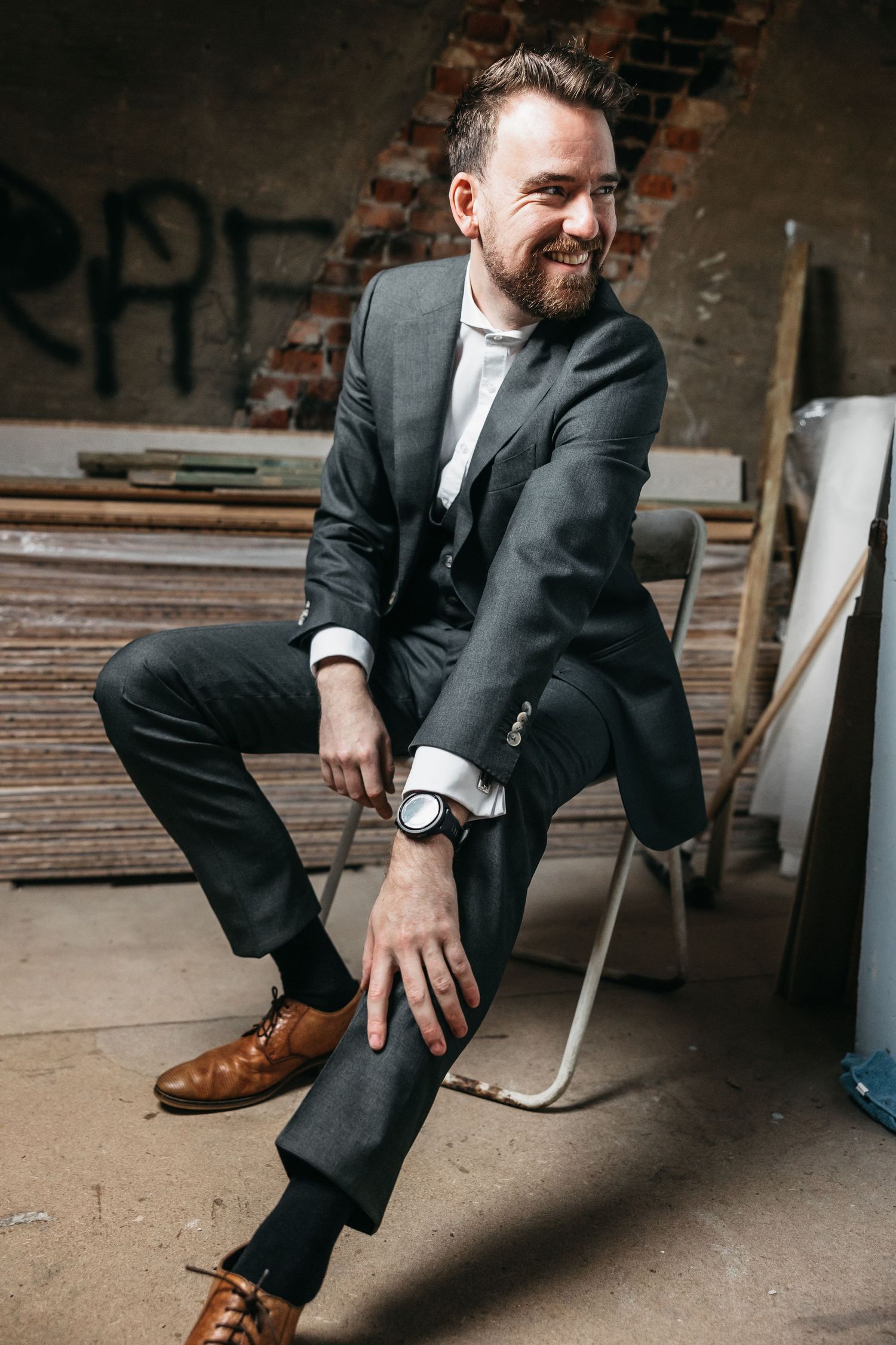 Smiling bearded groom in gray three-piece suit and brown shoes sitting in rustic venue.