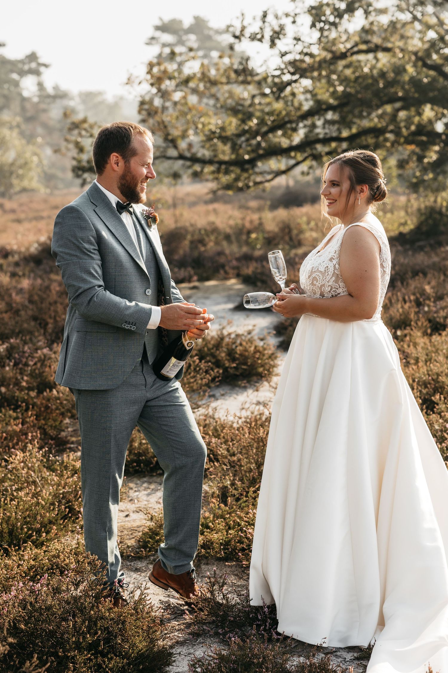 Wedding couple sharing champagne in sunlit heathland, bride in white gown and groom in gray suit.